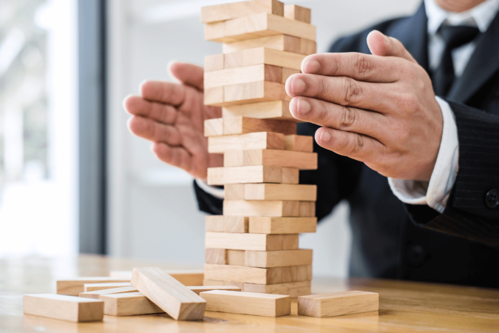L'image montre une personne en costume jouant à un jeu de Jenga. L'individu utilise ses deux mains pour stabiliser ou retirer un bloc de la tour en bois partiellement démontée. Plusieurs blocs sont déjà sur la table, indiquant que le jeu est en cours. Cette scène symbolise la planification minutieuse et stratégique nécessaire pour maintenir la stabilité et éviter l'effondrement, tout comme il est crucial de garantir la continuité et la résilience des services IT pendant les perturbations. Un plan de reprise d'activité dans le cadre d'un service IT doit être soigneusement élaboré pour restaurer les systèmes critiques, protéger les données sensibles et minimiser les interruptions de service, assurant ainsi la continuité des opérations de l'entreprise.