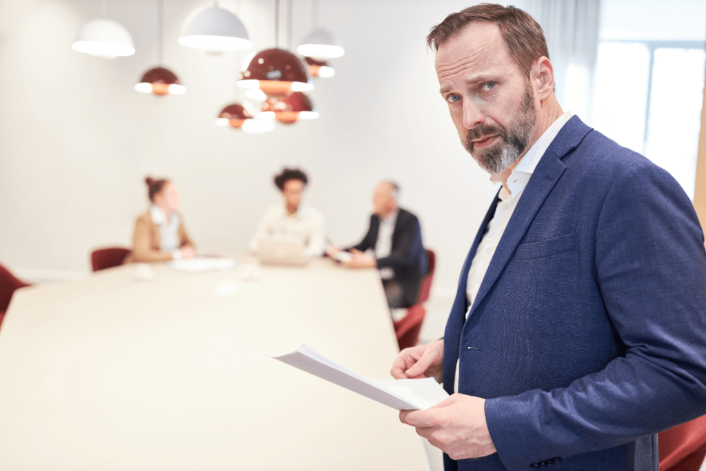 Au premier plan, une personne en costume bleu examine attentivement des documents, symbolisant la préparation et la stratégie. En arrière-plan, trois autres personnes sont assises autour d'une grande table de réunion, certaines avec des ordinateurs portables. Cette scène illustre les préoccupations des dirigeants concernant l'externalisation du service IT. La présence d'ordinateurs portables représente les risques liés à la confidentialité des données sensibles, tandis que la discussion animée entre les participants reflète la crainte de perdre le contrôle sur des processus stratégiques et la baisse de réactivité face aux urgences techniques. La salle bien éclairée met en avant le caractère sensible de ces discussions.