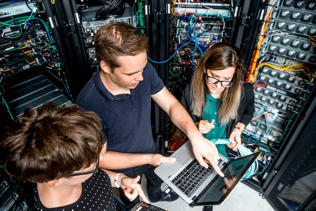 Trois spécialistes IT travaillant dans un centre de données, entourés de serveurs et d'équipements réseau. L'un des experts utilise un ordinateur portable et pointe vers l'écran, tandis qu'un autre manipule un câble réseau. Cette scène illustre l'importance essentielle du service IT pour les PME modernes. En gérant et en maintenant les infrastructures technologiques, ces professionnels assurent la continuité des opérations, la sécurité des données et l'optimisation des systèmes. Leur rôle est indispensable pour permettre aux PME de rester compétitives et efficaces dans un monde numérique en constante évolution.